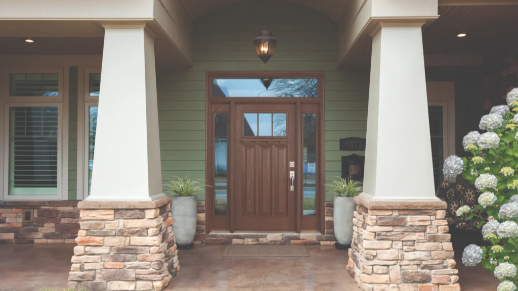 Front porch of a house with green siding, stone-clad columns, and a dark wooden front door with two potted plants nearby.