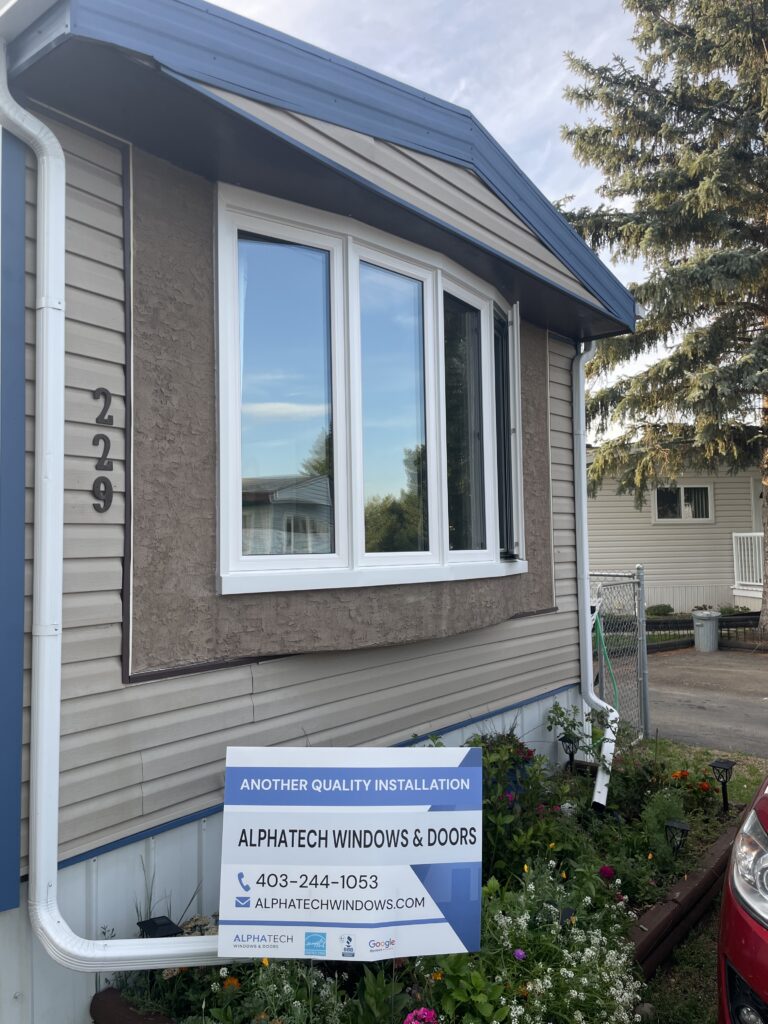 White-framed bay window on a beige house with a blue metal awning; sign for Alphatech Windows & Doors in front.