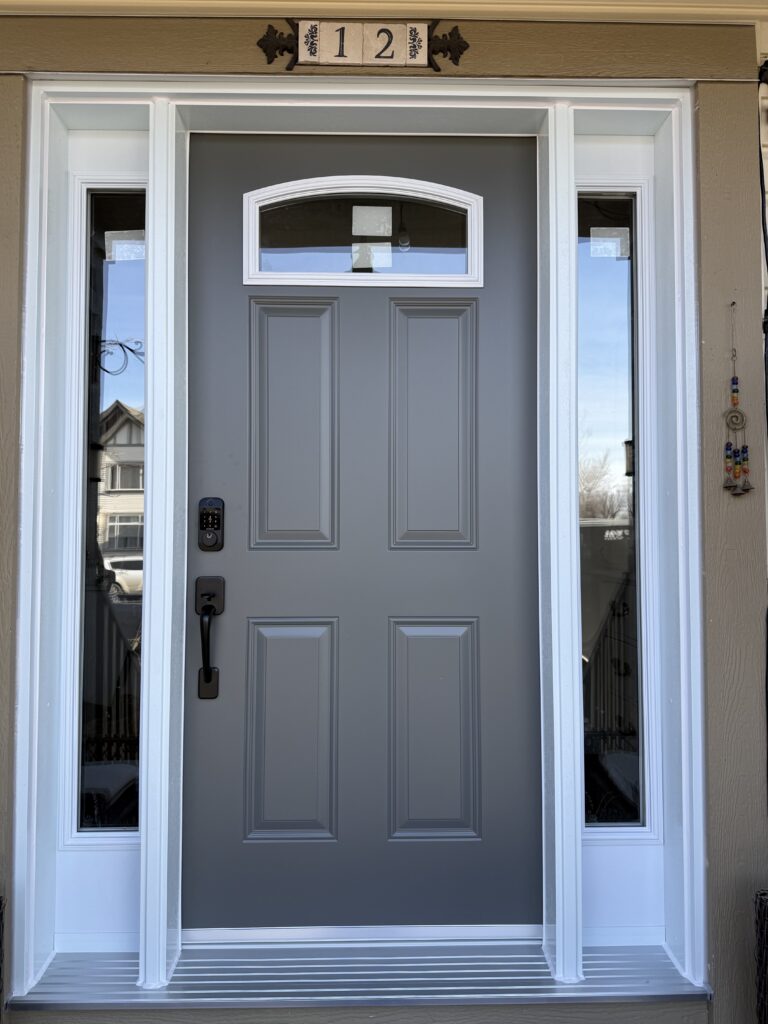 Gray front entry door with raised panels, flanked by white-trimmed glass sidelights and a decorative '12' plaque above the door.