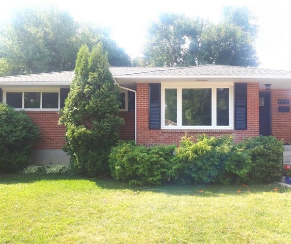 Red brick ranch-style house with a large front window framed by blue shutters, a tall evergreen shrub, and trimmed hedges in a sunny front yard.